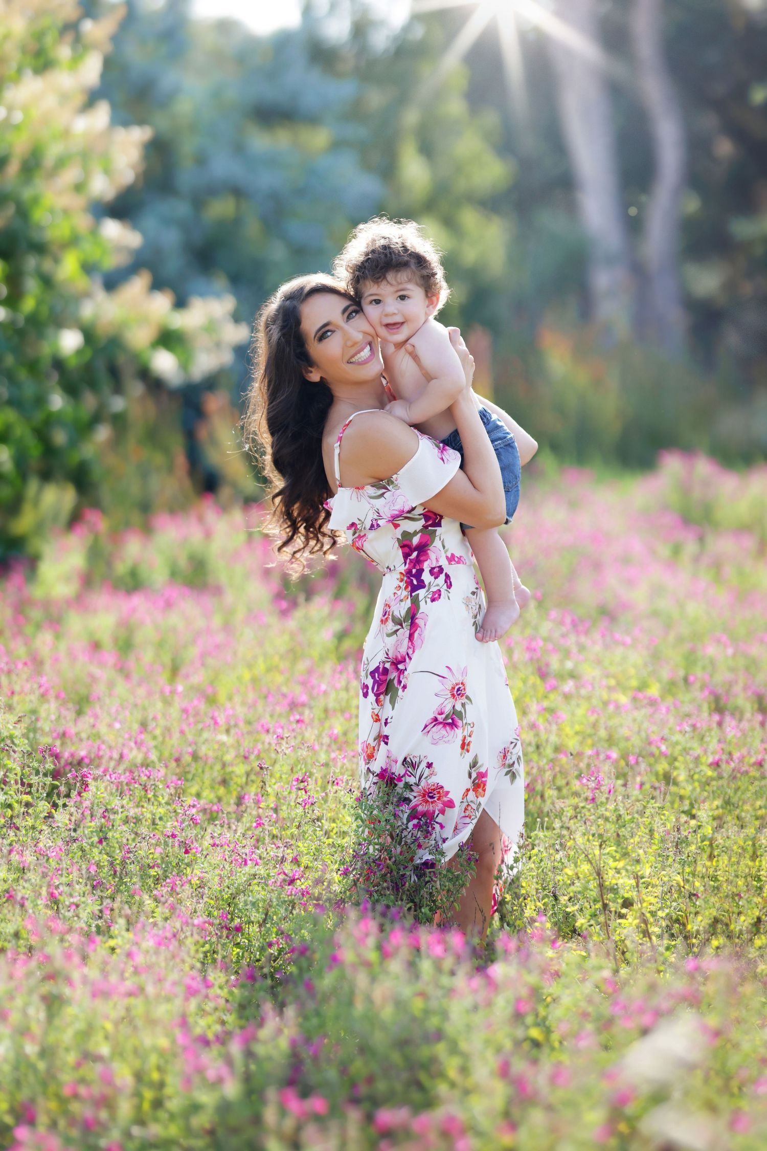 A smiling mother in a floral dress holds her toddler in a sunny pink wildflower field.