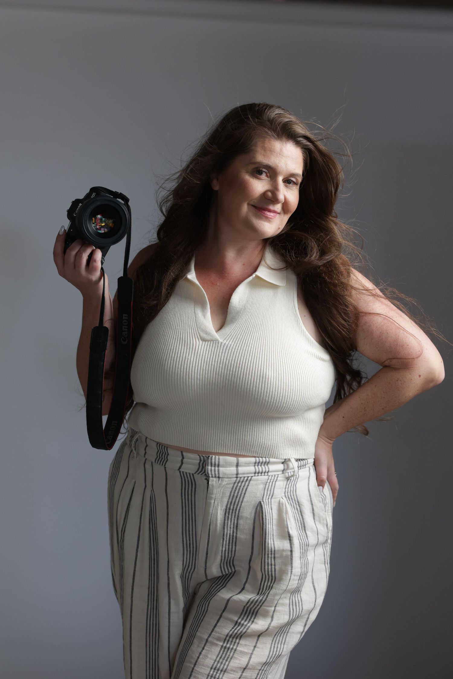 Smiling woman with long hair holding DSLR camera in gray background studio portrait.