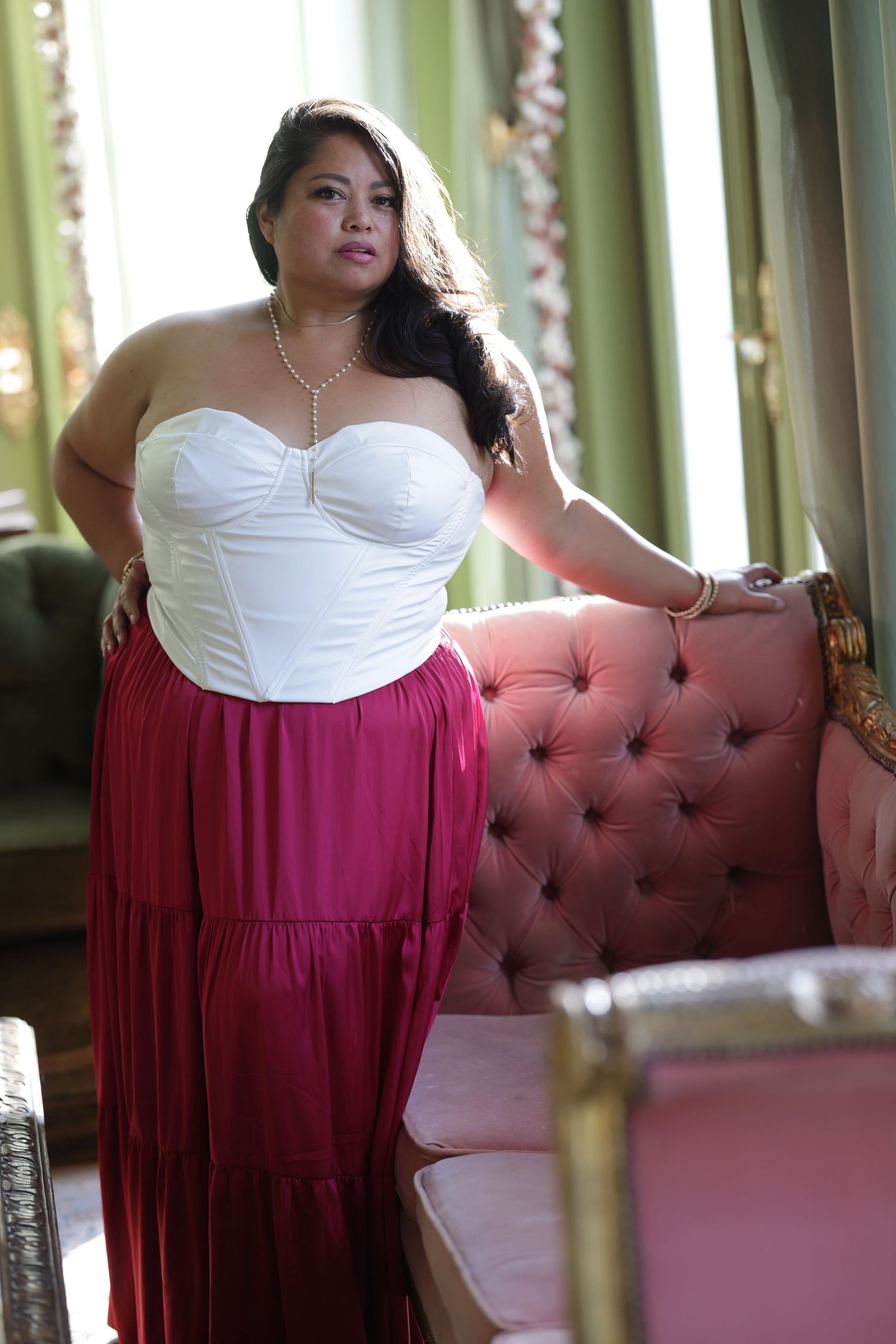 Plus-size woman in white strapless corset top and magenta maxi skirt poses elegantly beside a pink tufted sofa.