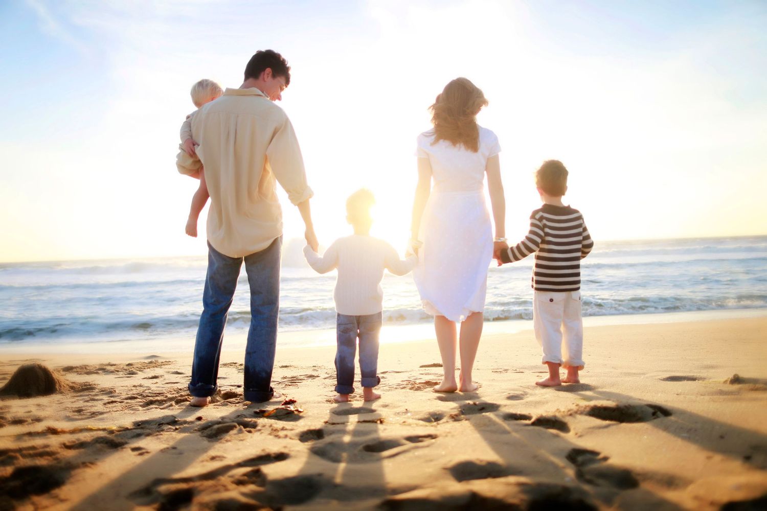 Family of five holding hands on a sunny beach, viewed from behind, with ocean waves and golden sunset light.