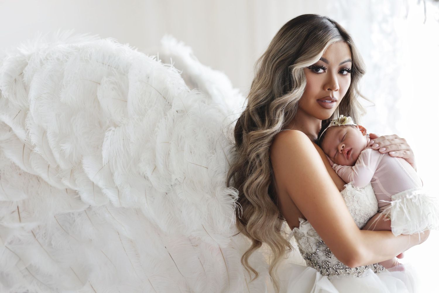 Woman with angel wings holding a sleeping newborn baby in a dreamy white studio portrait.
