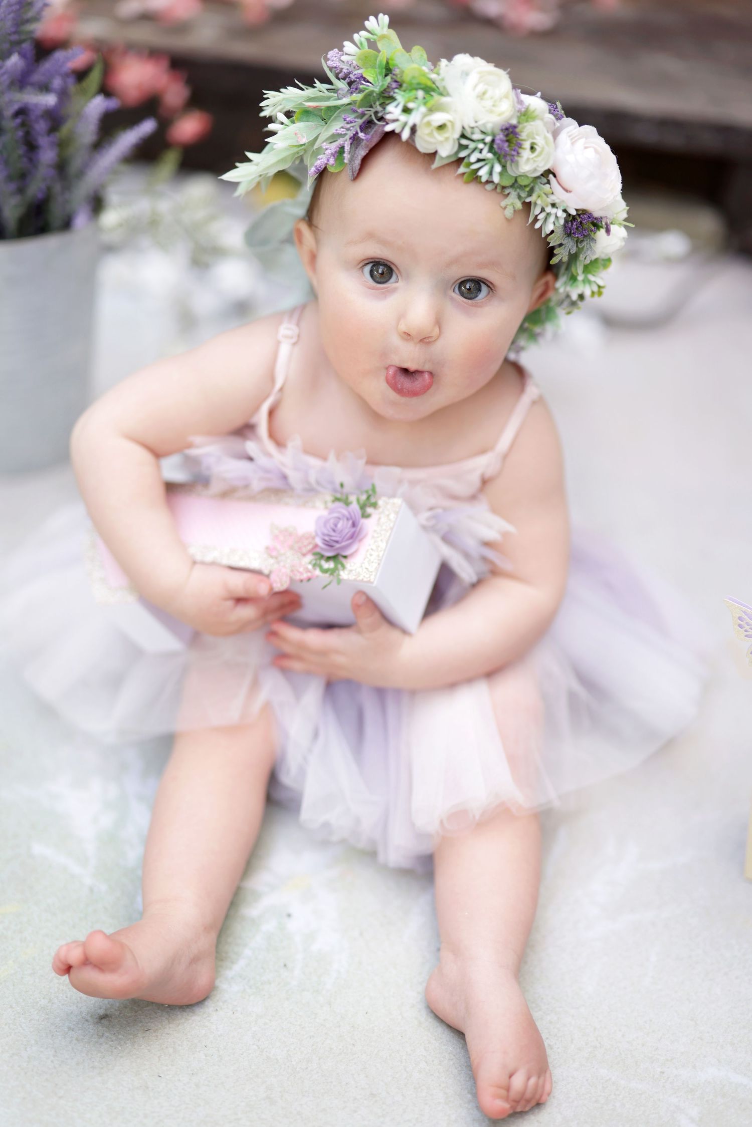 Adorable baby girl in lavender tutu dress and floral crown holds a gift box while sitting on the floor.
