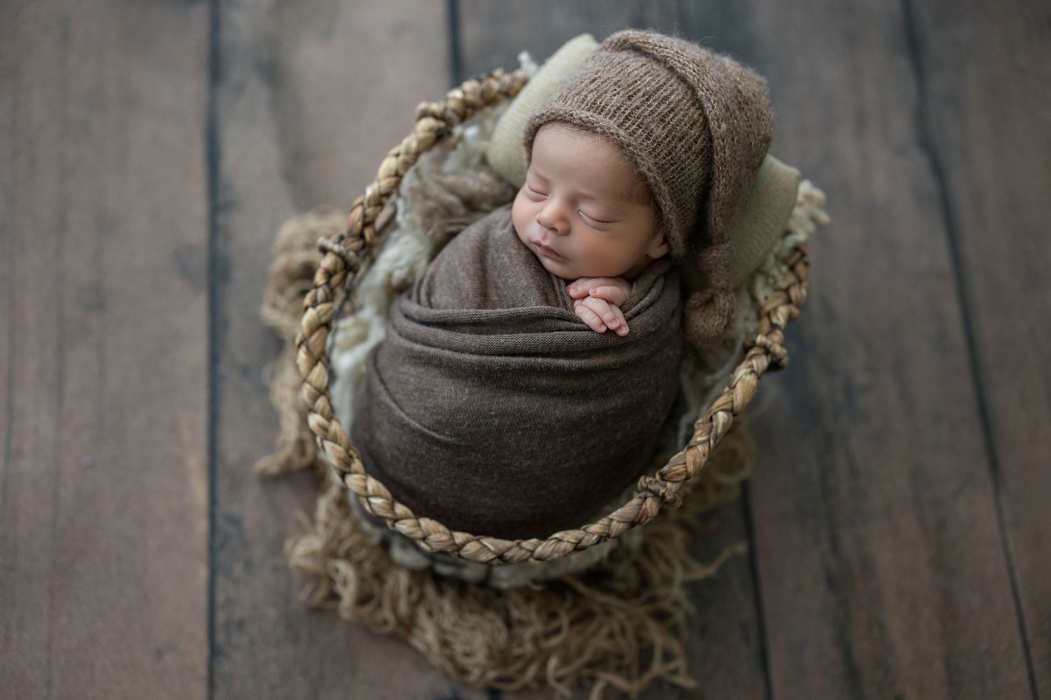 Sleeping newborn baby swaddled in brown wrap wearing knit bonnet nestled in a woven basket on wood floor.