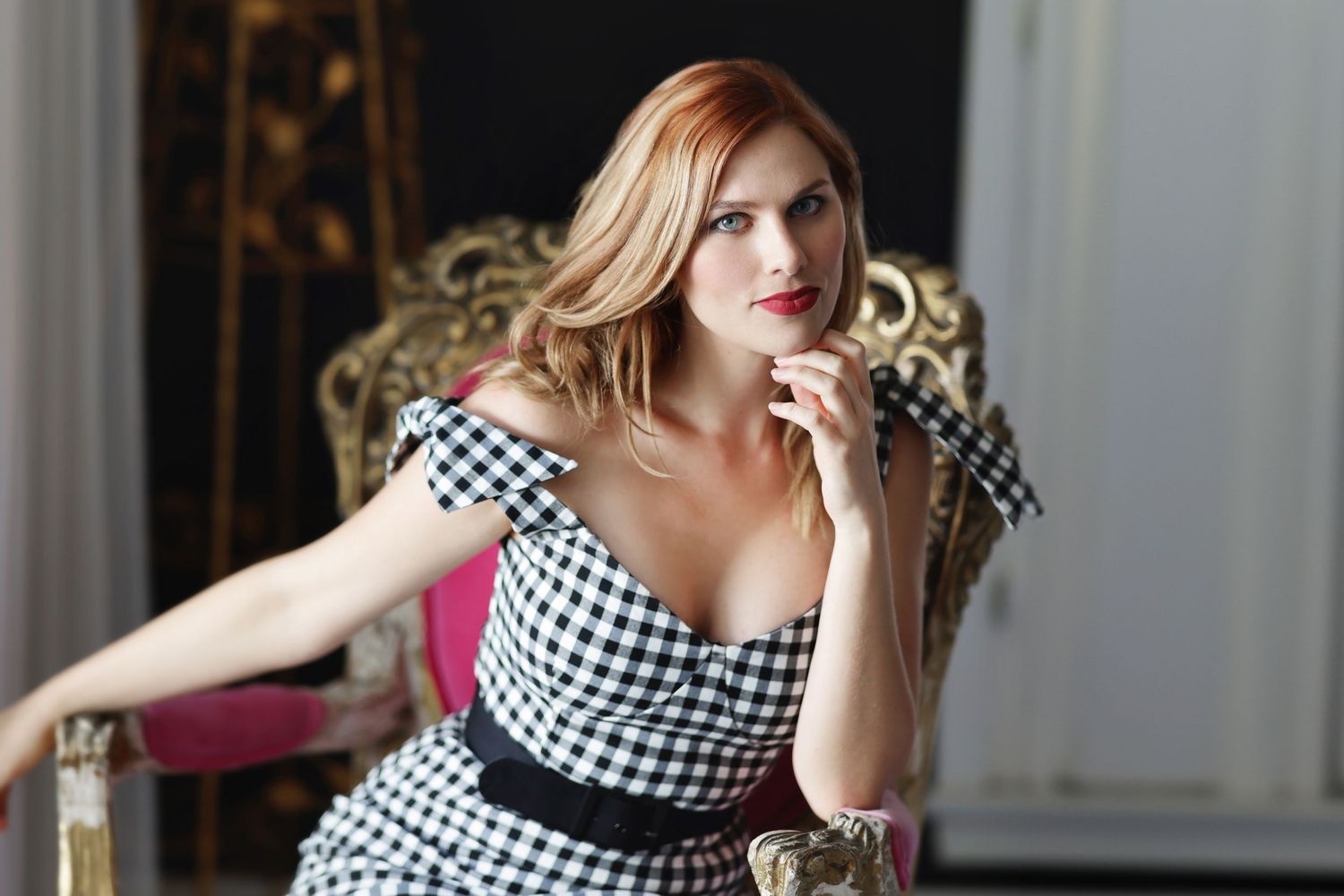 Elegant redhead woman in checkered off-shoulder dress seated in ornate gold chair, posing gracefully.