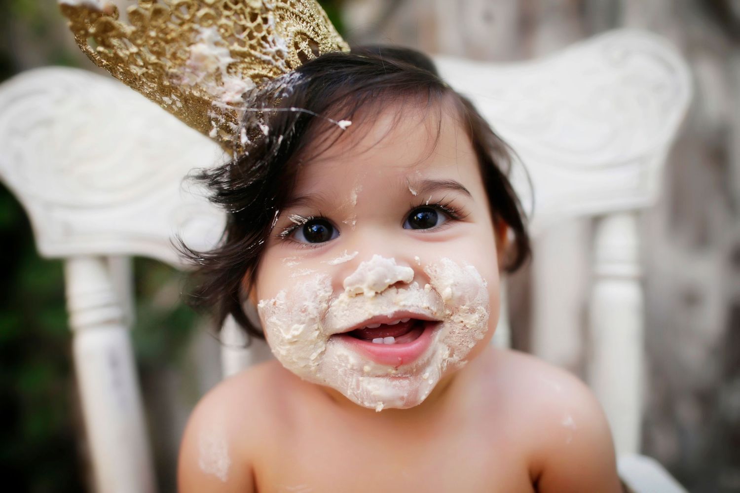Happy baby with cake smeared face wearing gold crown smiles at first birthday cake smash.