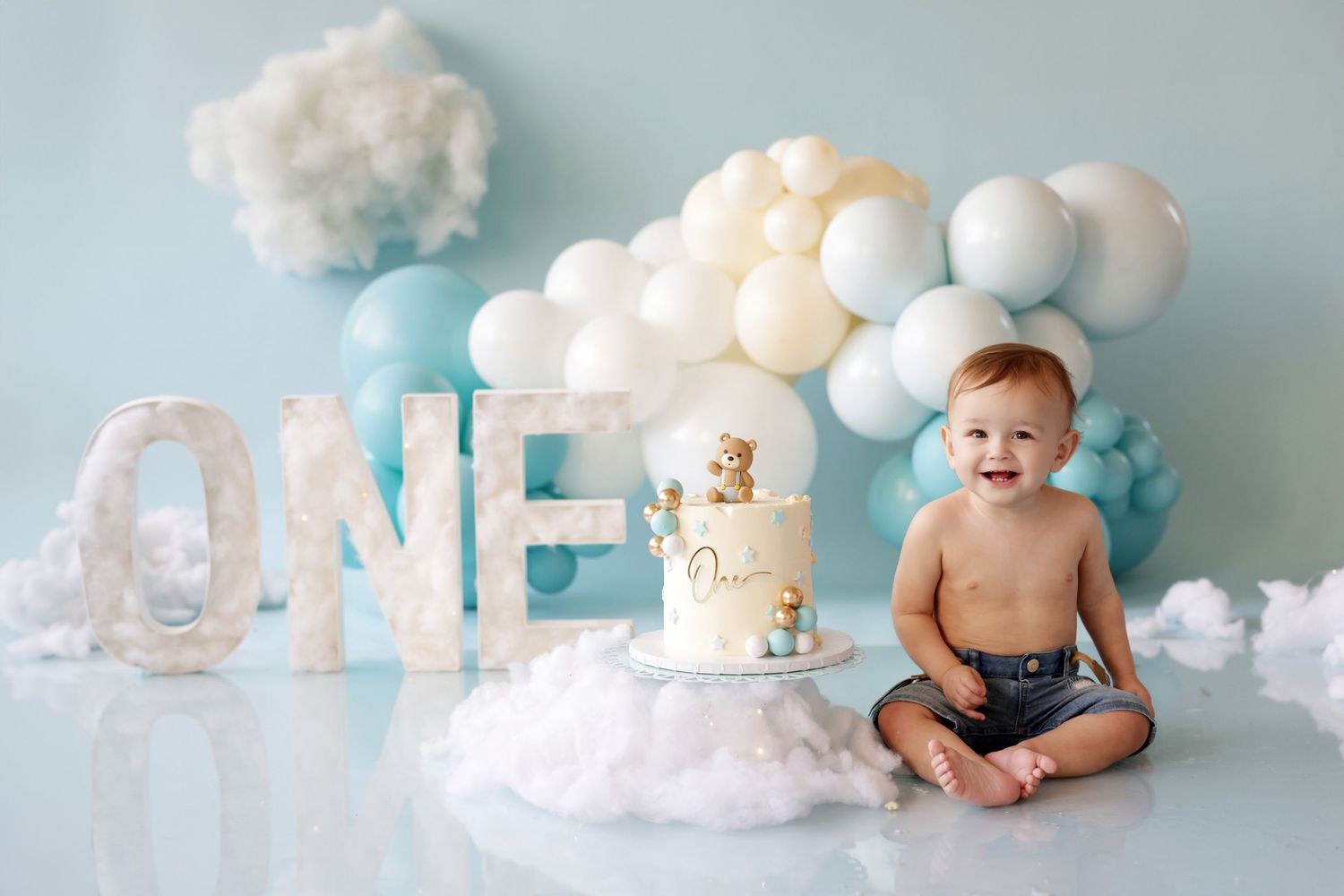 Laughing baby boy in denim shorts celebrating first birthday with ONE letters and balloon backdrop.