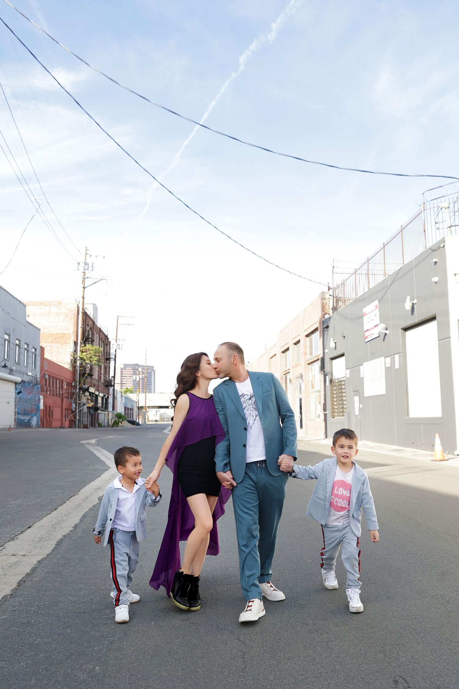 A family of four walks down an urban street, parents kissing while holding hands with their two young sons.