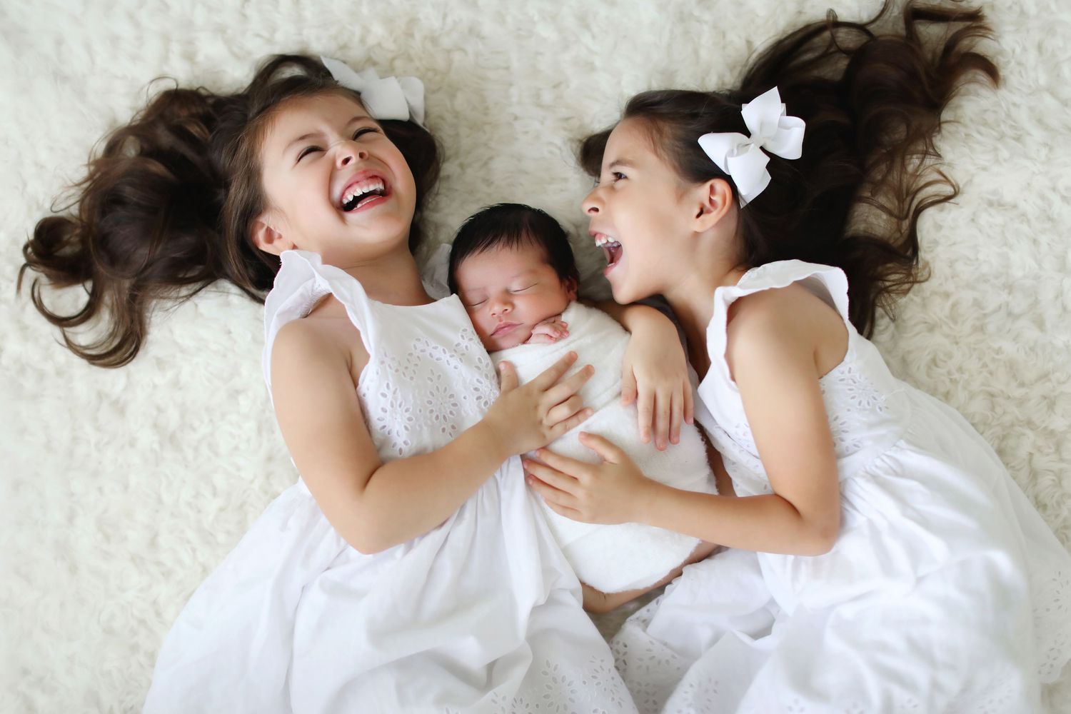Two young girls in white dresses laughing while lying beside a sleeping newborn baby on a white fluffy rug.
