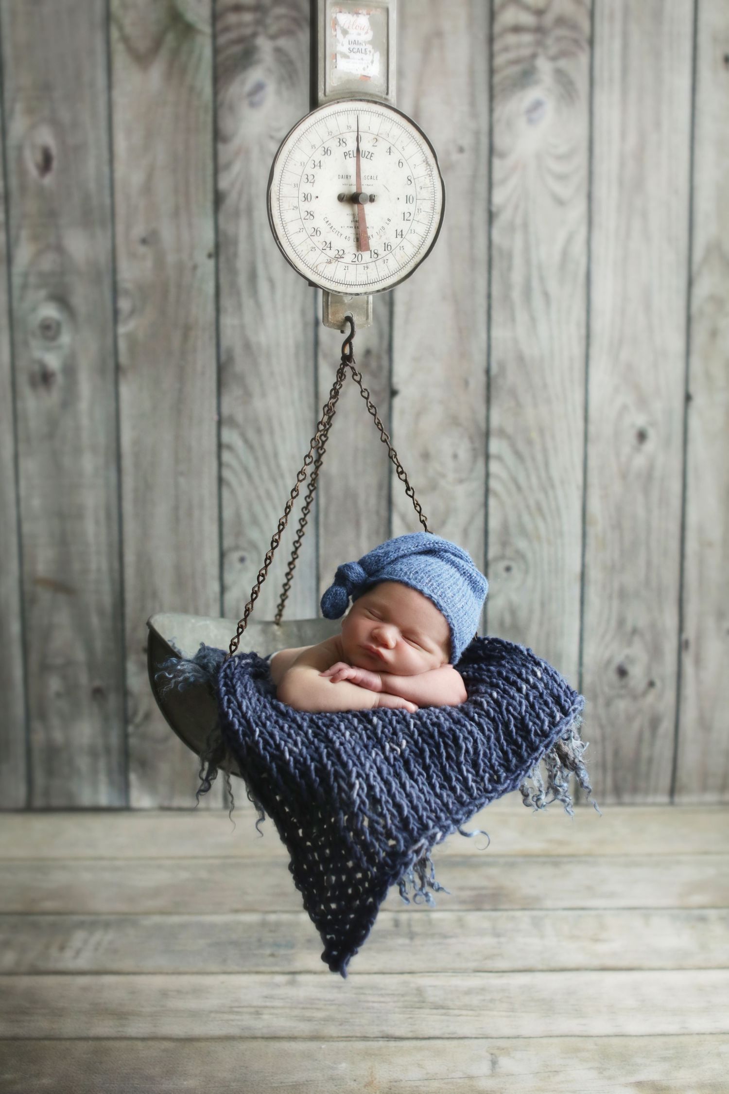 Sleeping newborn baby wearing a blue knit hat resting on a vintage hanging scale with a navy blanket.