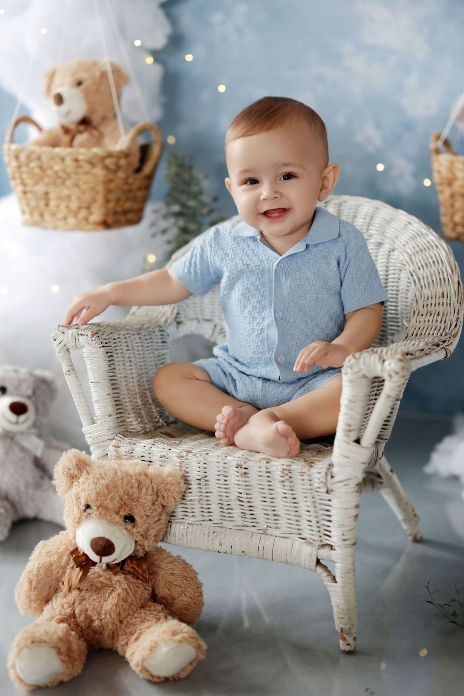 Smiling baby boy in blue romper sitting in white wicker chair with teddy bears in a whimsical holiday nursery.