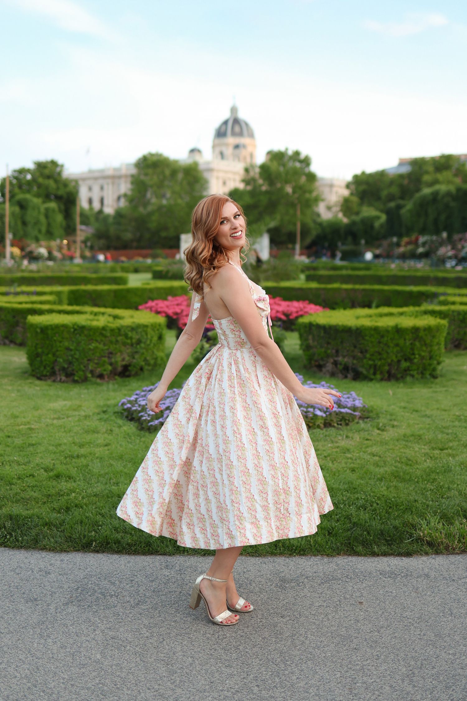 Smiling redhead woman in floral midi dress twirling in formal garden near historic building.