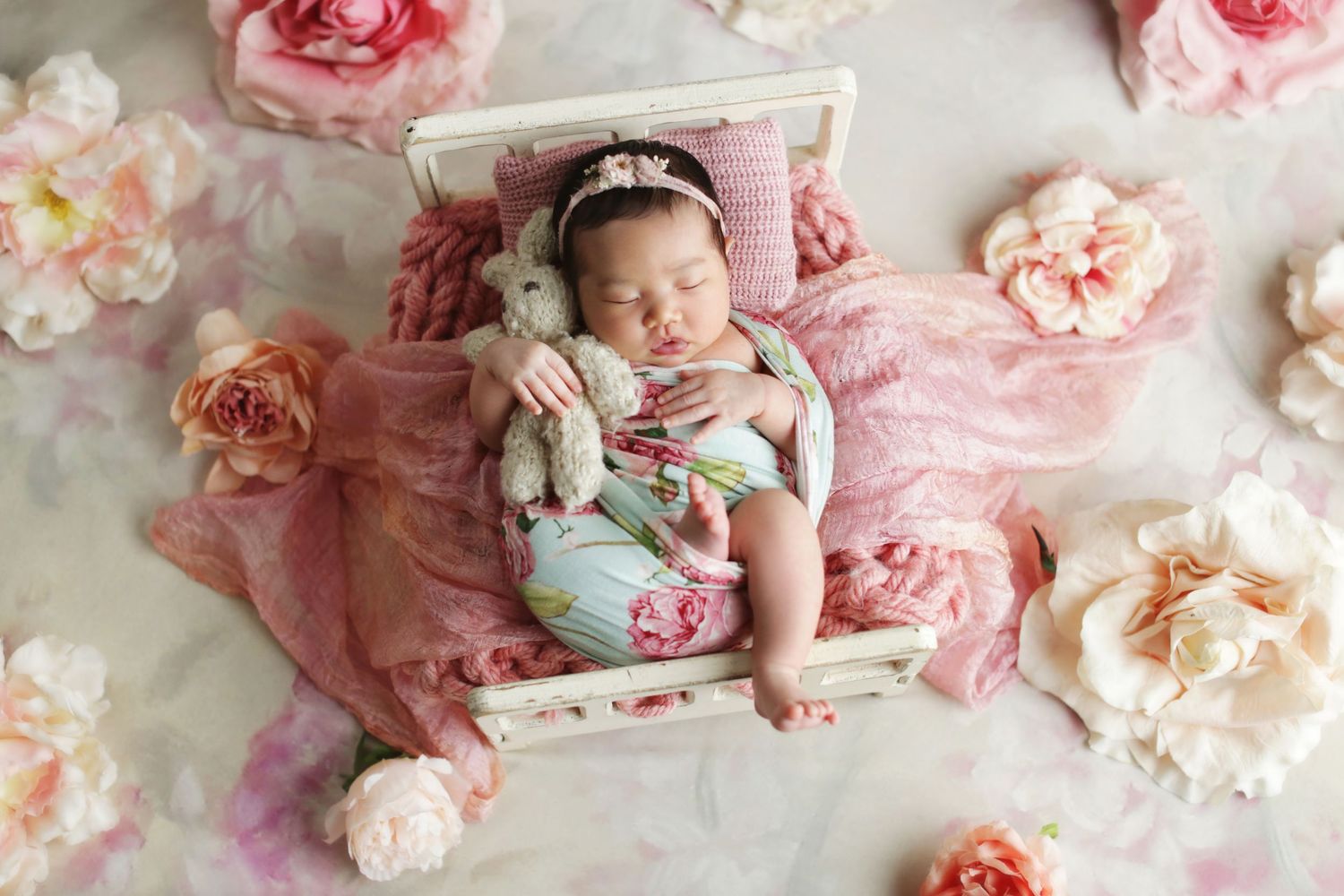 Newborn baby girl sleeping peacefully in a tiny white bed with a stuffed bunny, surrounded by pink roses.