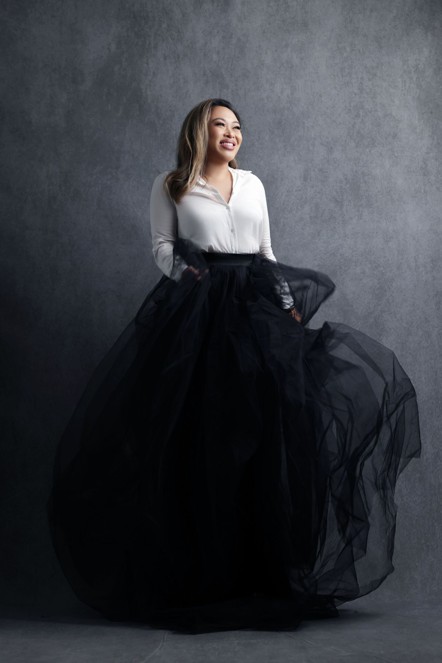Woman in white blouse and flowing black tulle skirt smiling elegantly in a professional studio portrait.