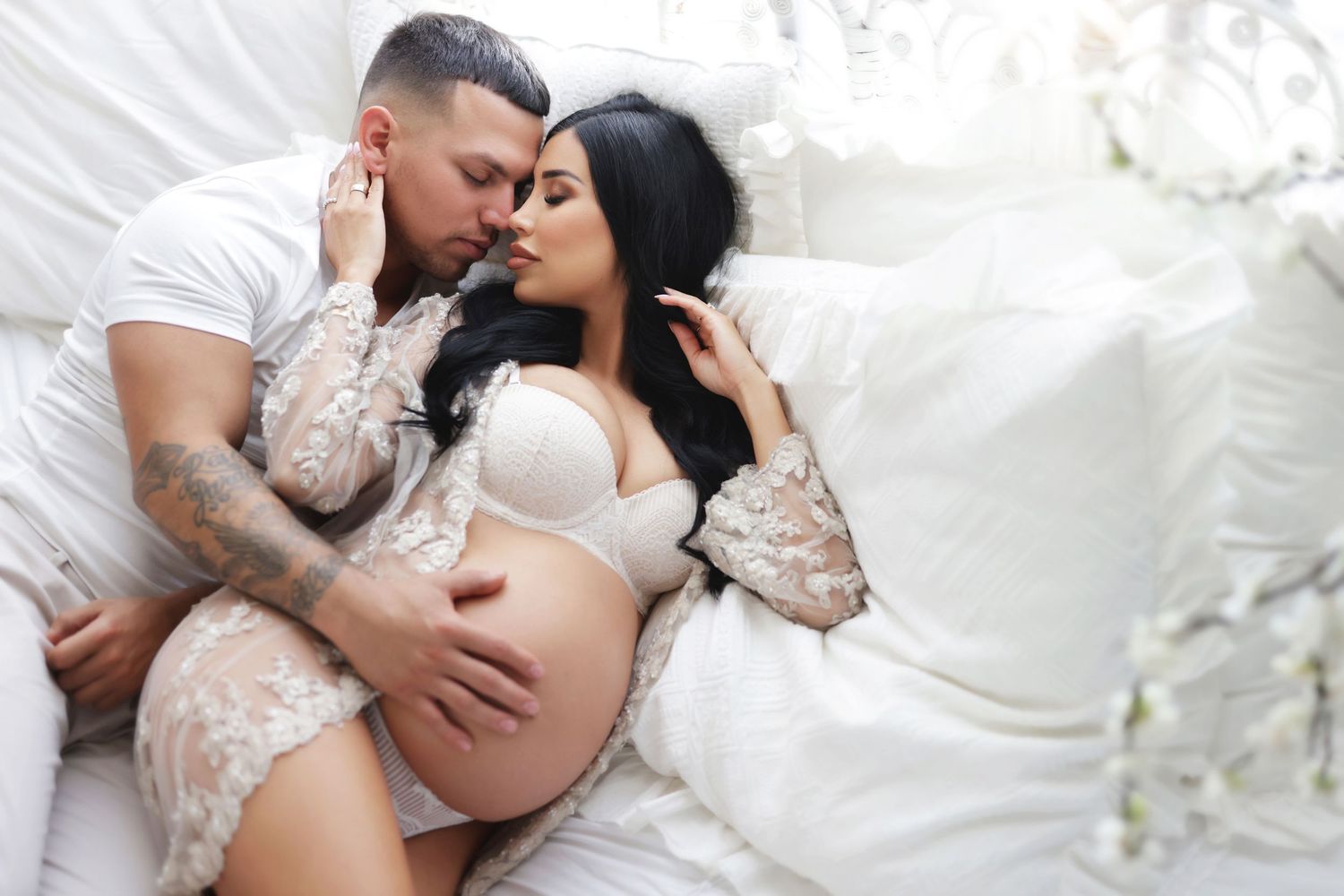 A tattooed man tenderly kisses a pregnant woman in white lace lingerie while cradling her bare baby bump on a white bed.