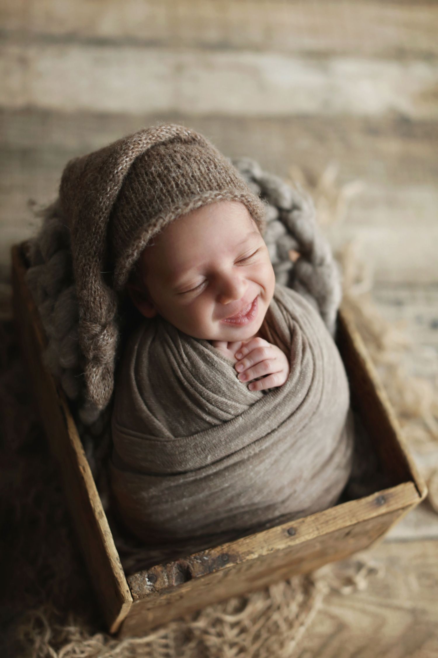 Smiling swaddled newborn in knit hat nestled in rustic wooden crate.