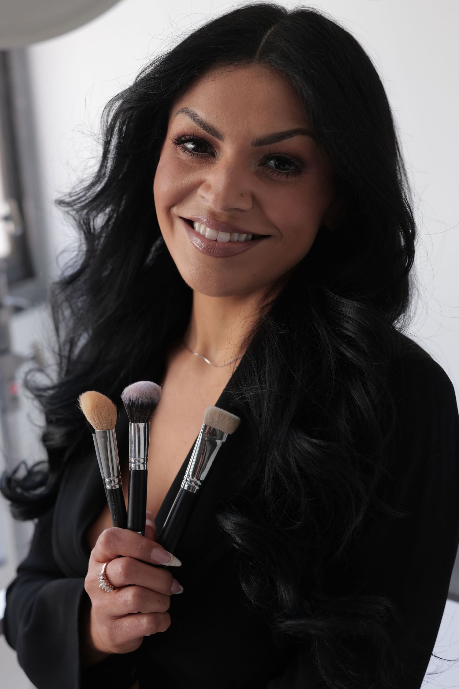 Smiling makeup artist with long dark hair holding professional brushes in black blazer.