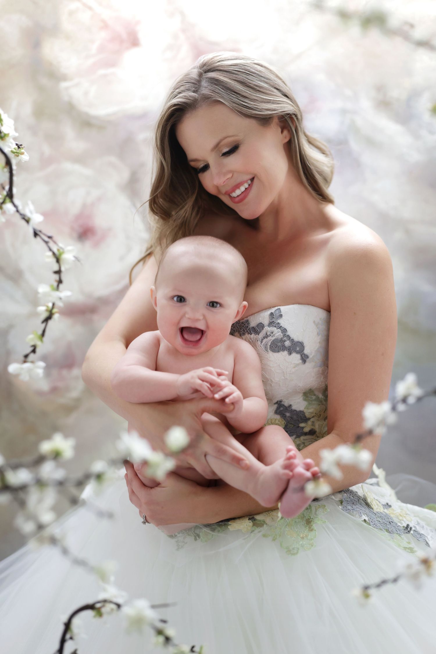 Smiling woman in white floral gown holding a laughing baby surrounded by spring cherry blossoms.