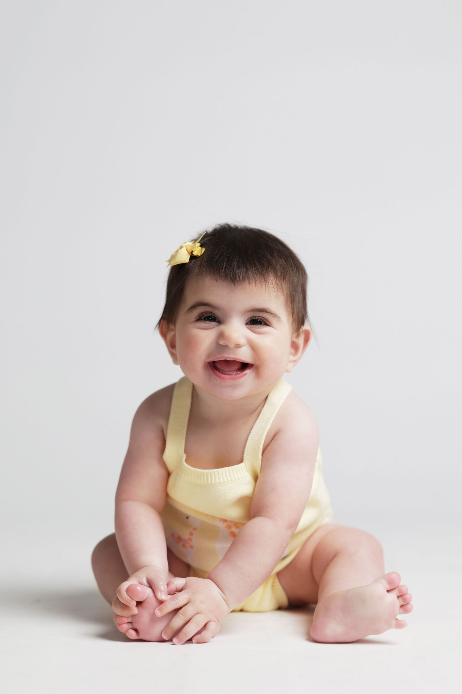 A happy baby girl in a yellow romper with a bow in her hair sits smiling on a white background.