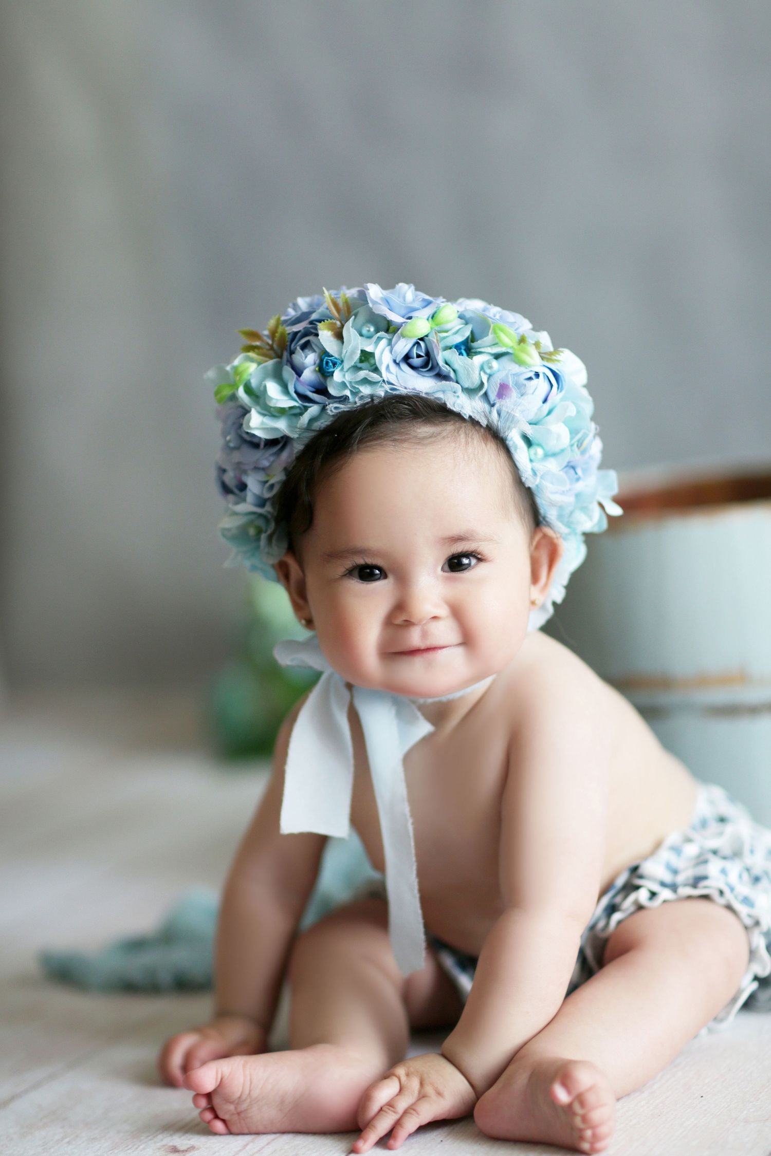 Adorable baby girl wearing a blue hydrangea floral bonnet, sitting and smiling in a soft, light-colored studio setting.