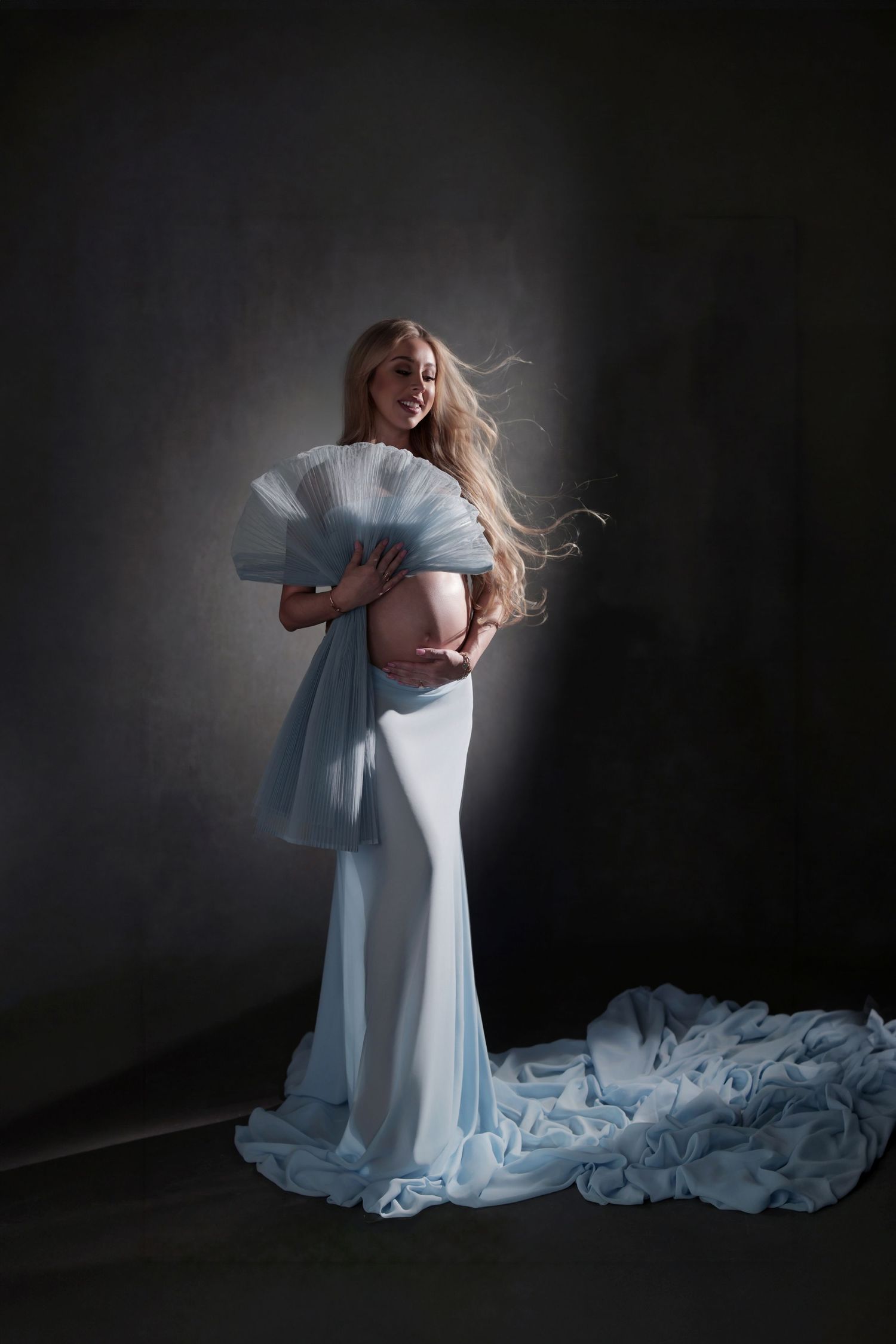 Smiling pregnant woman in elegant light blue gown with ruffled train.