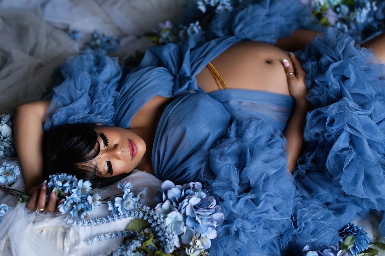 Pregnant woman in blue tulle gown lying among blue hydrangeas for a dreamy maternity photo shoot.