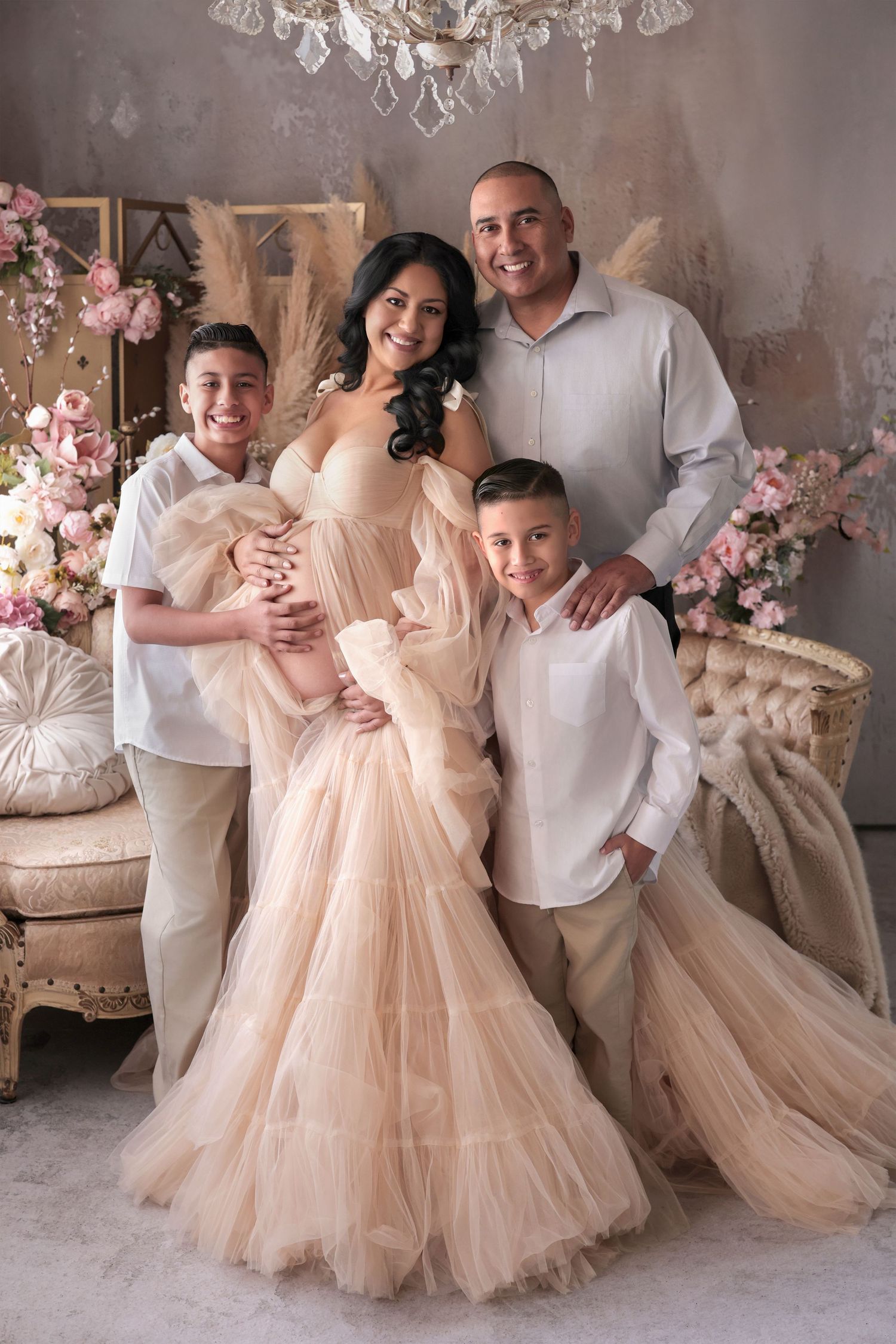 Family maternity photo shoot with pregnant woman in elegant champagne tulle gown, husband, and two sons in floral studio.