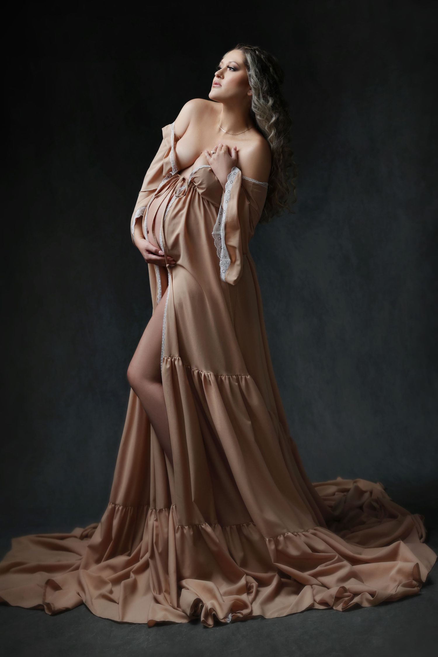 Pregnant woman in an elegant flowing champagne off-shoulder gown posing on a dark studio background.