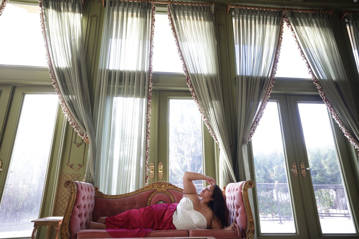 Woman in red maxi skirt reclines gracefully on ornate pink sofa beneath tall draped windows in elegant room.