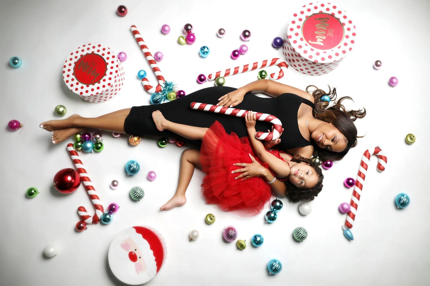 Two women lying on white floor surrounded by colorful Christmas ornaments, candy canes, and festive decorations.