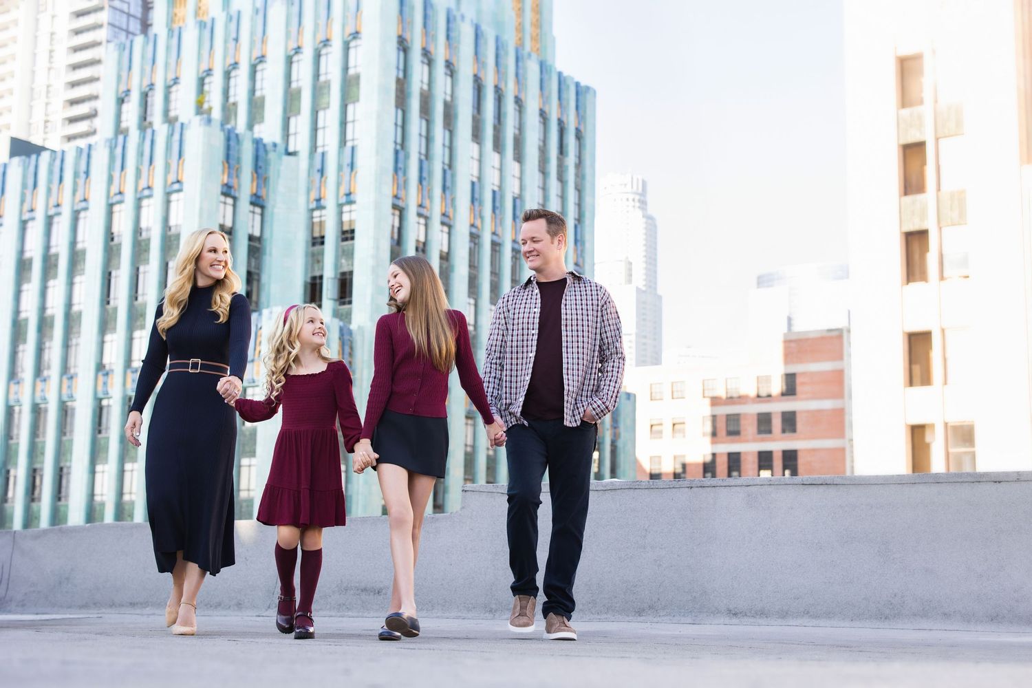 Family of four walking hand-in-hand outdoors with modern city buildings in the background on a sunny day.