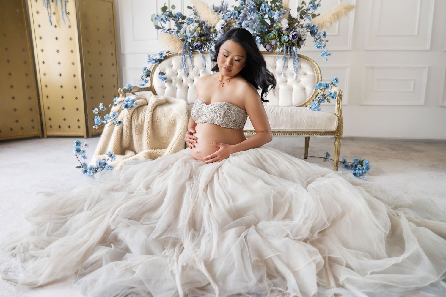 Pregnant woman in elegant tulle gown sits gracefully on ornate sofa surrounded by blue floral decorations.