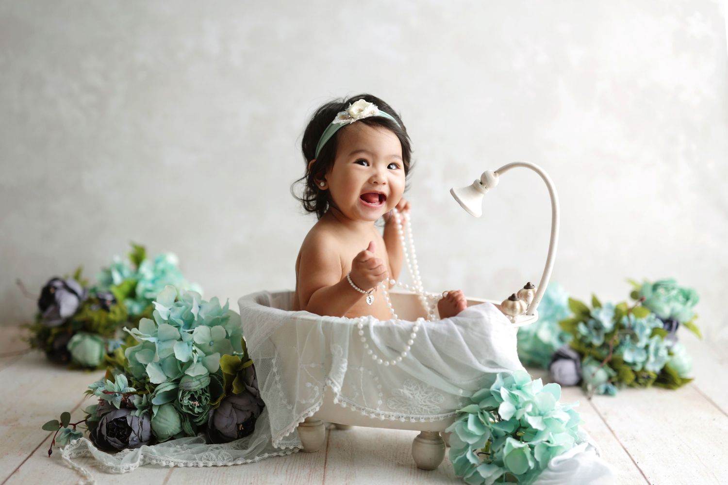Happy baby girl sitting in a decorative bathtub with teal flowers and pearls.