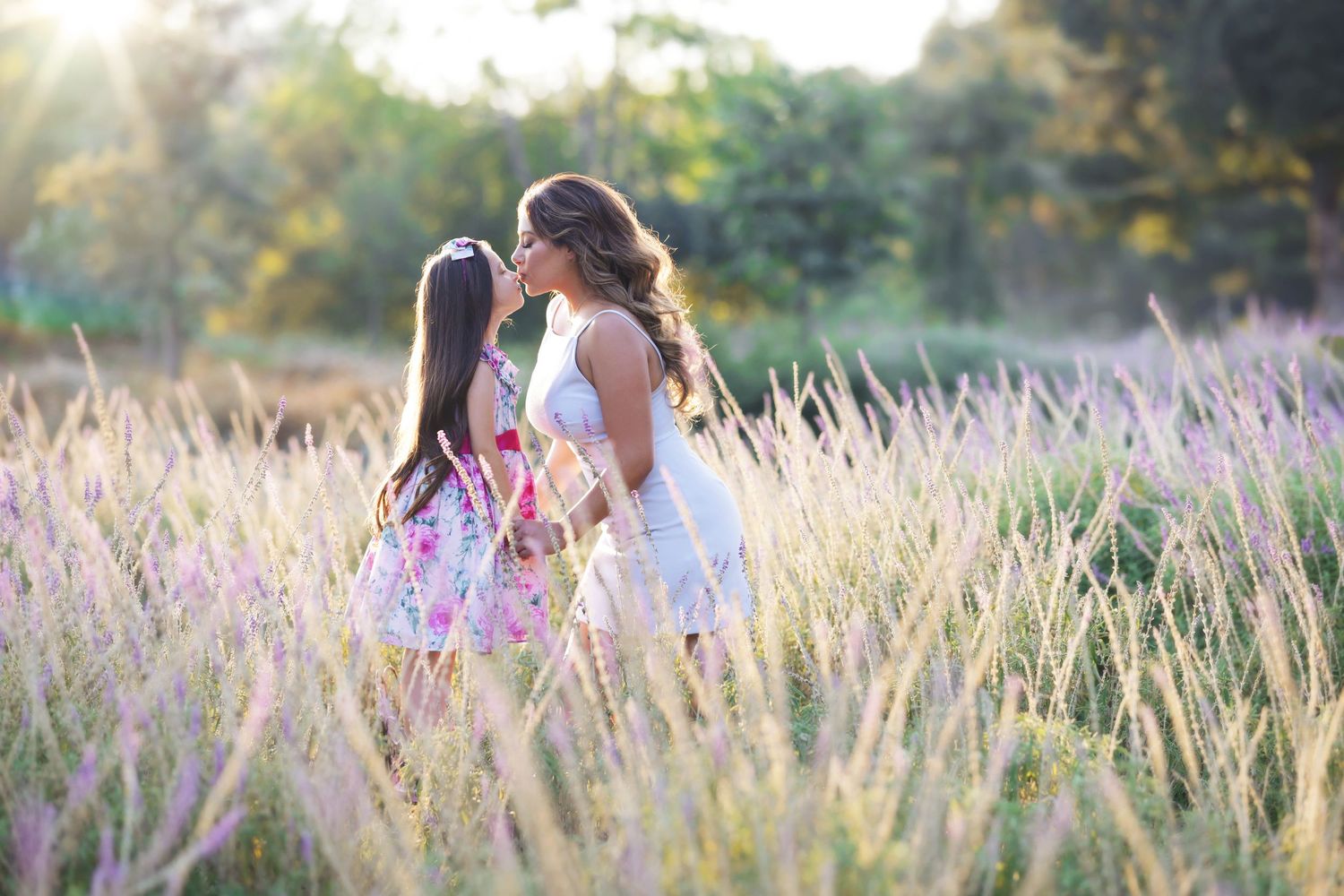 Two women in floral dresses walk through a sunlit field of tall pink wildflowers during golden hour.