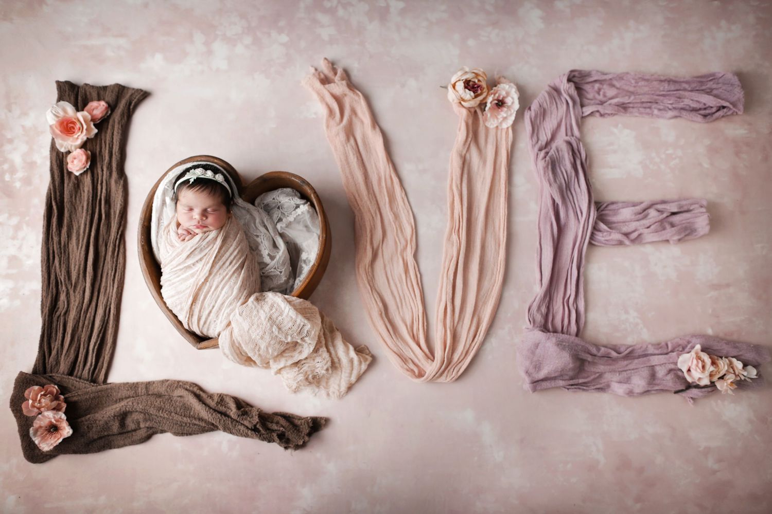 Newborn baby wrapped in white swaddle posed in heart bowl surrounded by fabric spelling LOVE with pink floral accents.