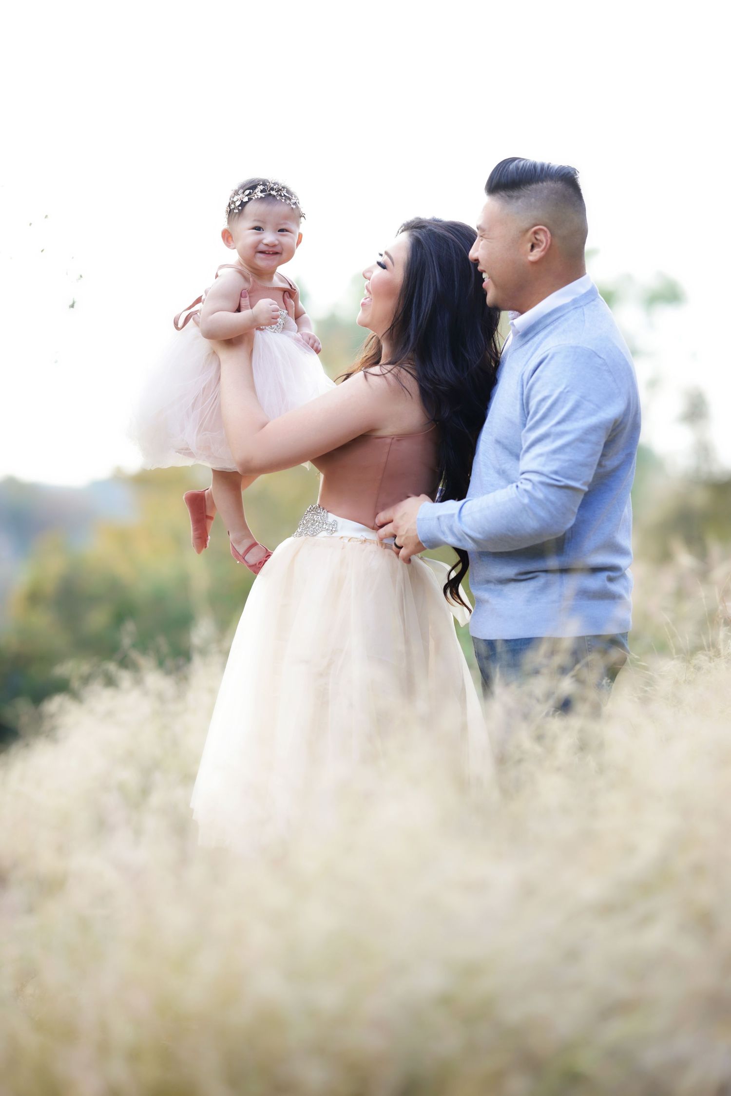 Happy family of three posing in golden field, mother lifting smiling baby girl in pink tutu dress.