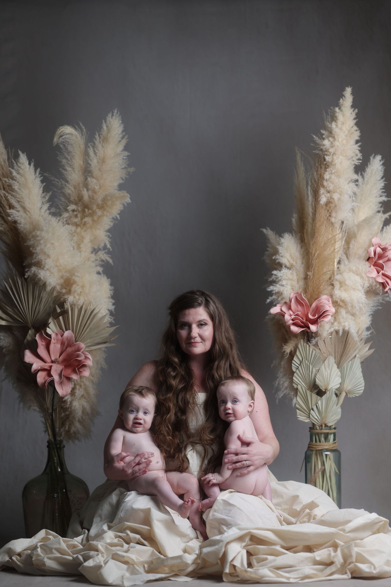 Mother with long brown hair holding twin babies surrounded by pampas grass and pink flowers.