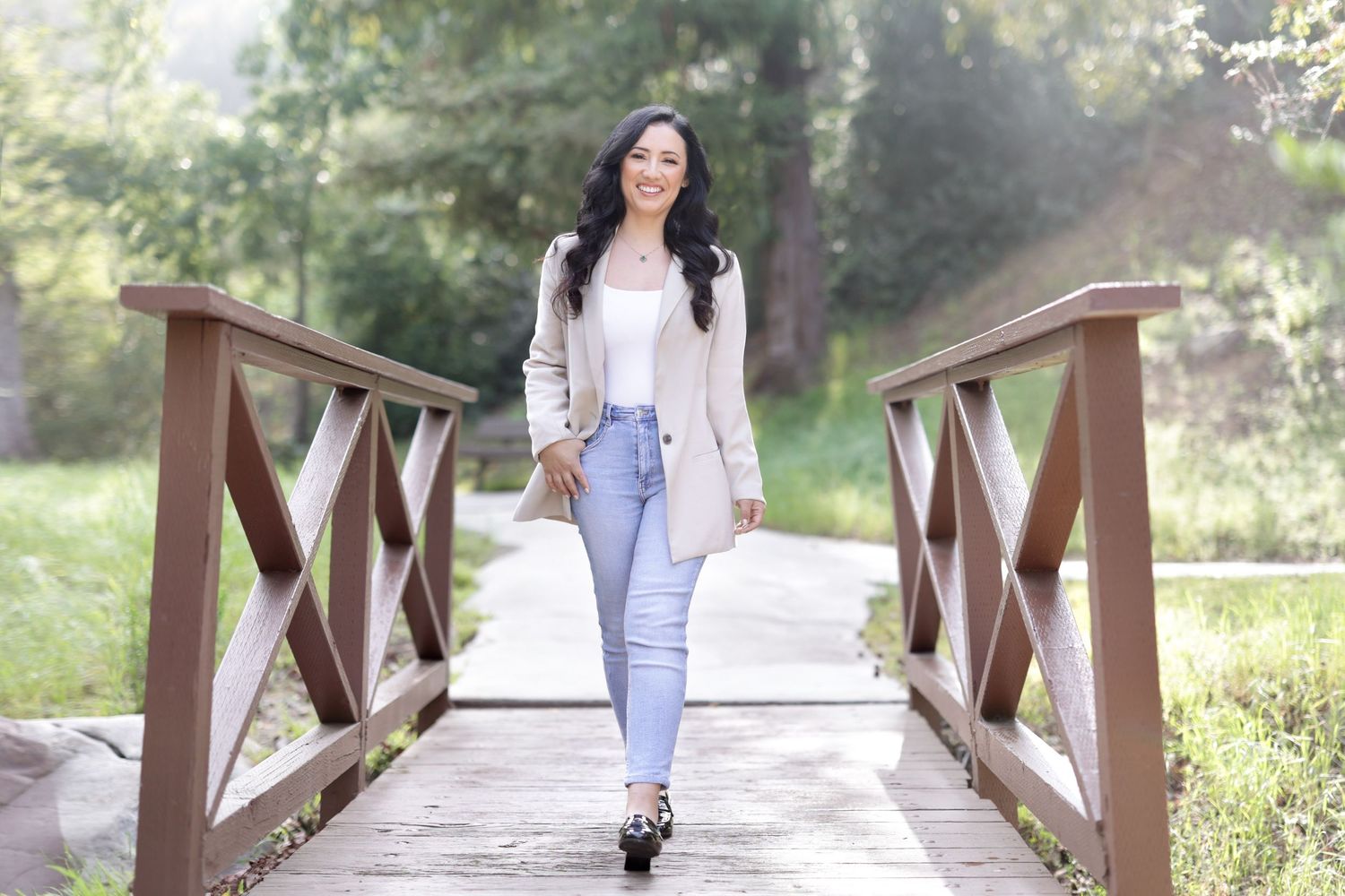 Woman in light blazer and jeans walking confidently on a wooden bridge in a lush green park.