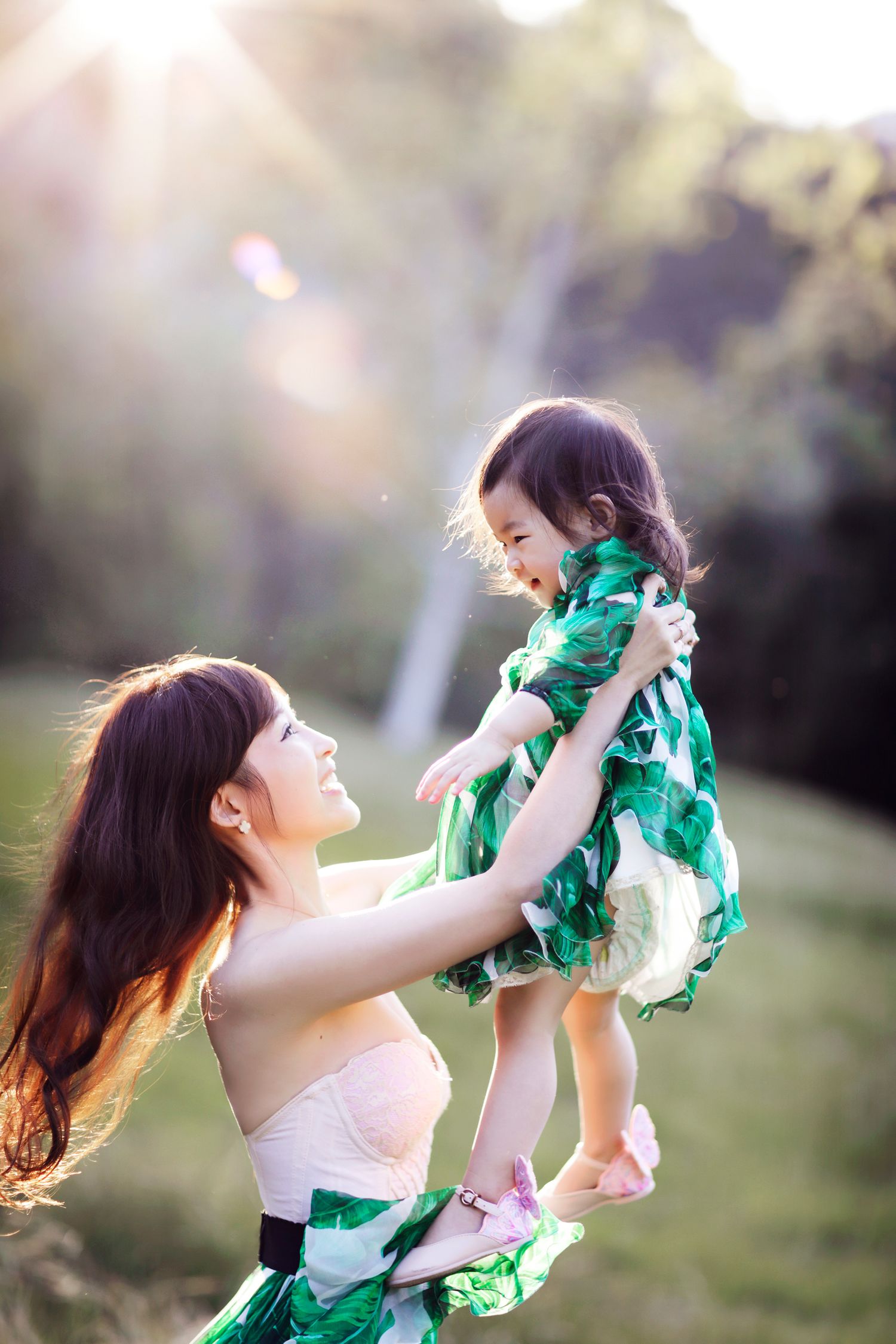 Mother lifting her smiling toddler daughter in a sunny park, both wearing matching green floral dresses.