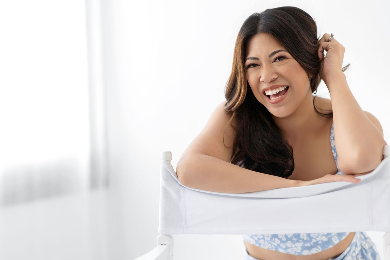 Smiling Asian woman with long dark hair posing casually in a bright white room wearing a blue floral top.