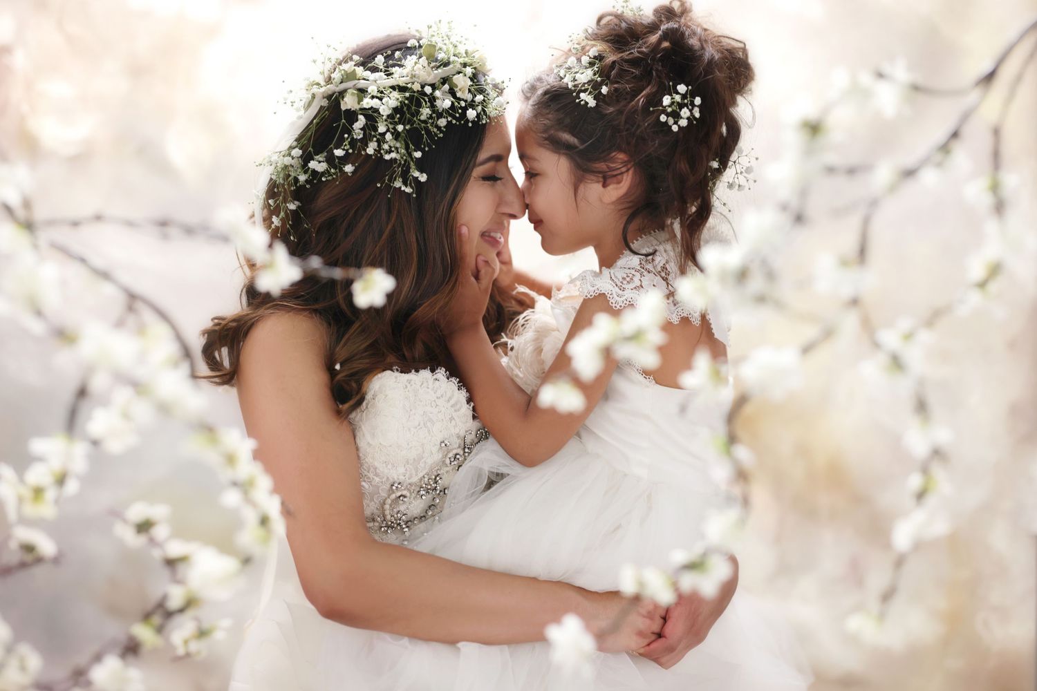 A mother and daughter in white dresses with floral crowns share a tender moment surrounded by spring blossoms.