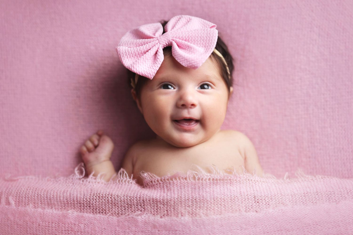 Adorable smiling baby girl wearing a pink bow headband lying on a soft pink blanket.