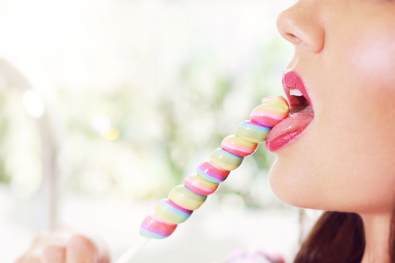 Close-up of a woman with pink lips licking a colorful spiral lollipop against a soft green background.