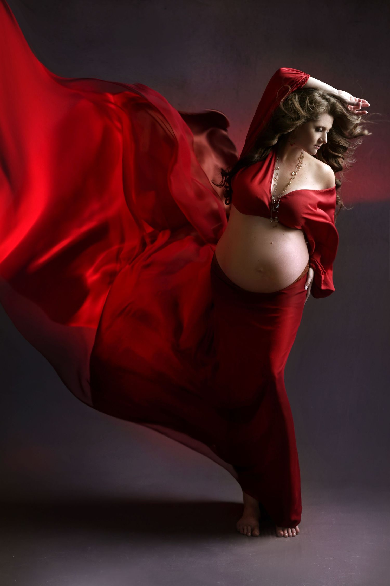 Pregnant woman in flowing red silk fabric dancing gracefully against a dark background.