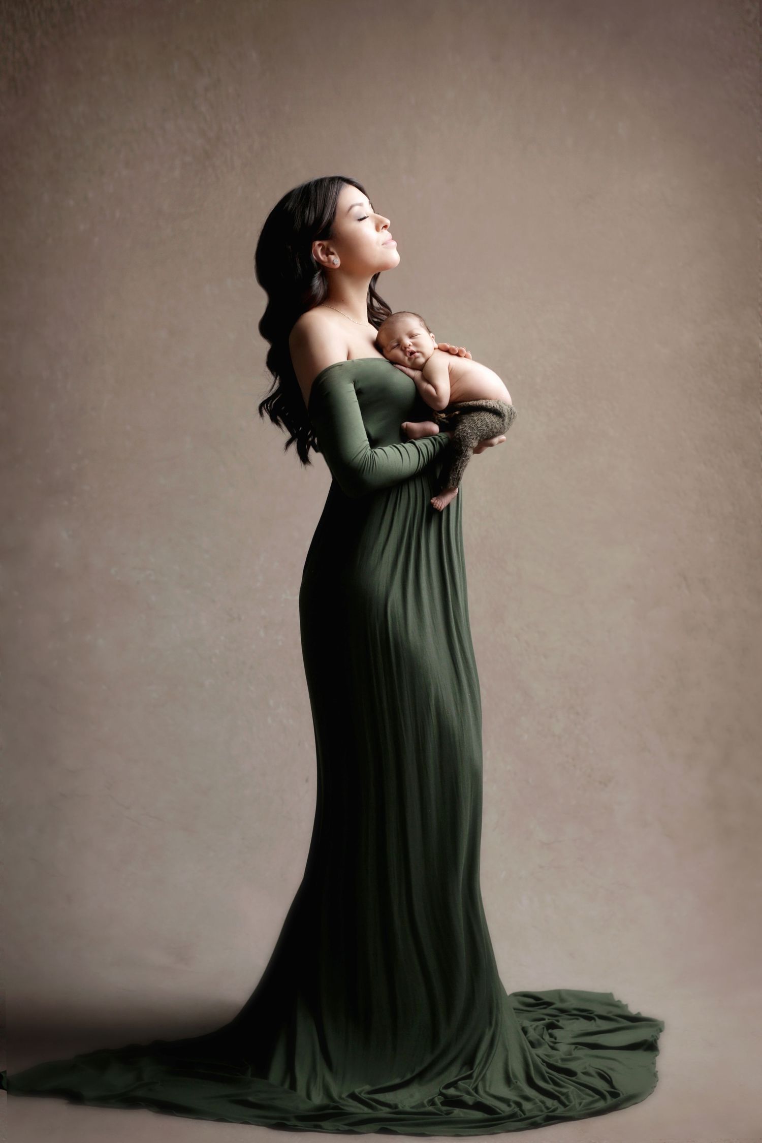 Woman in elegant green gown holding newborn baby in a studio portrait setting.