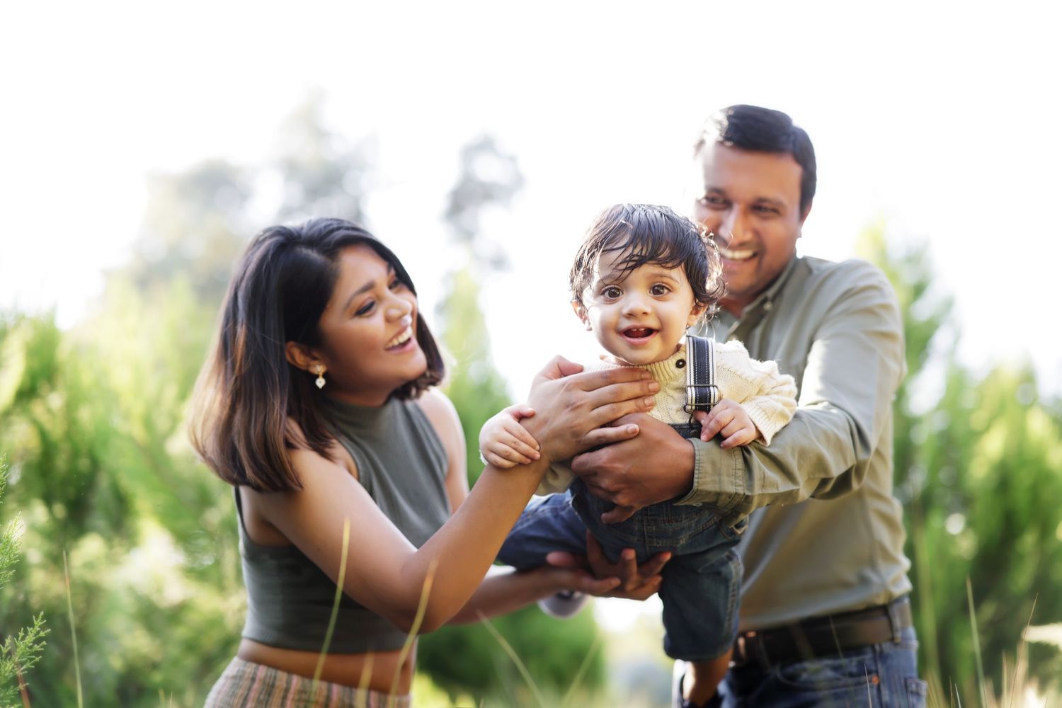 Happy Indian family with parents playing with toddler son outdoors in green park on sunny day.