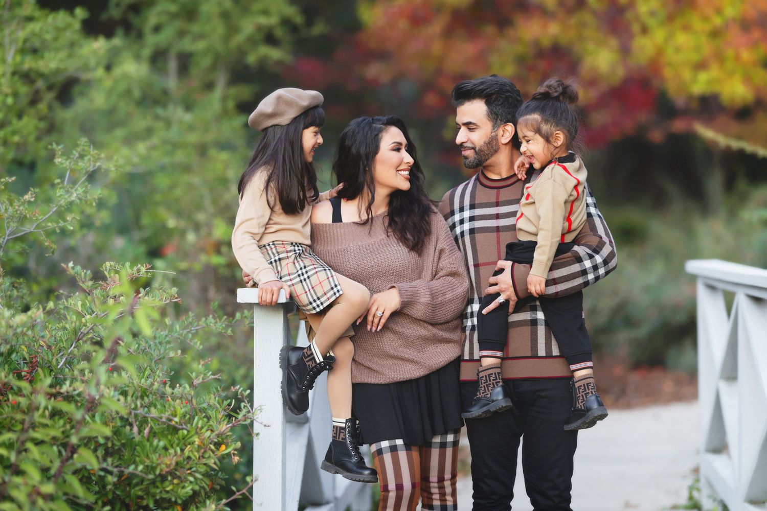 Happy family of four enjoying an autumn walk on a garden bridge, parents holding young children in plaid outfits.