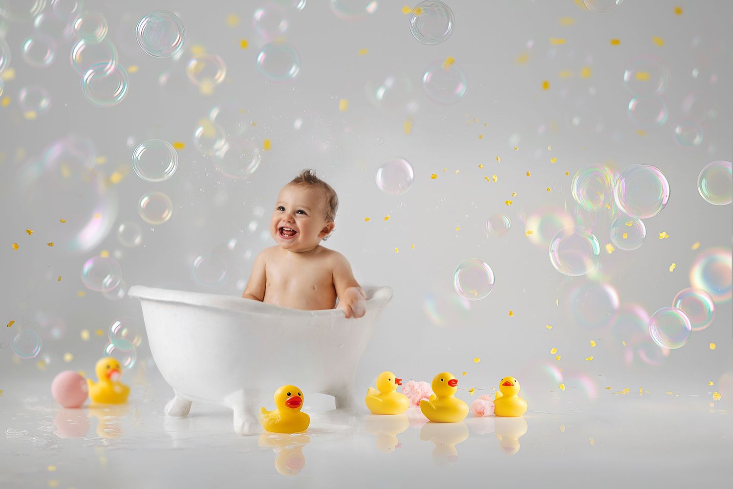 Happy baby laughing in white bathtub surrounded by rubber ducks and colorful soap bubbles on light background.