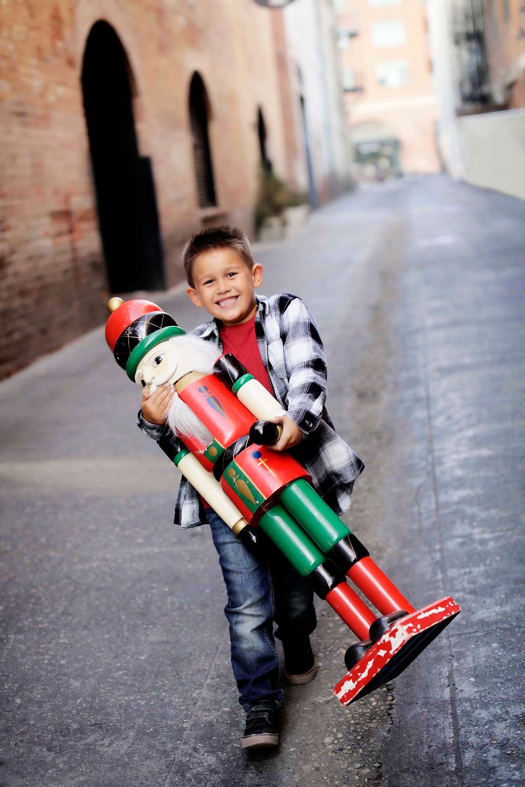 Smiling boy carrying a large Christmas nutcracker decoration while walking down an outdoor alley.