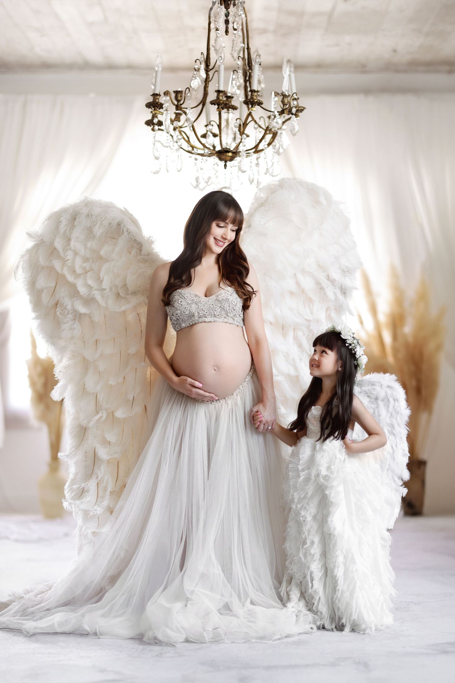 Pregnant woman and young girl dressed as angels in white gowns with feather wings pose in elegant white studio.