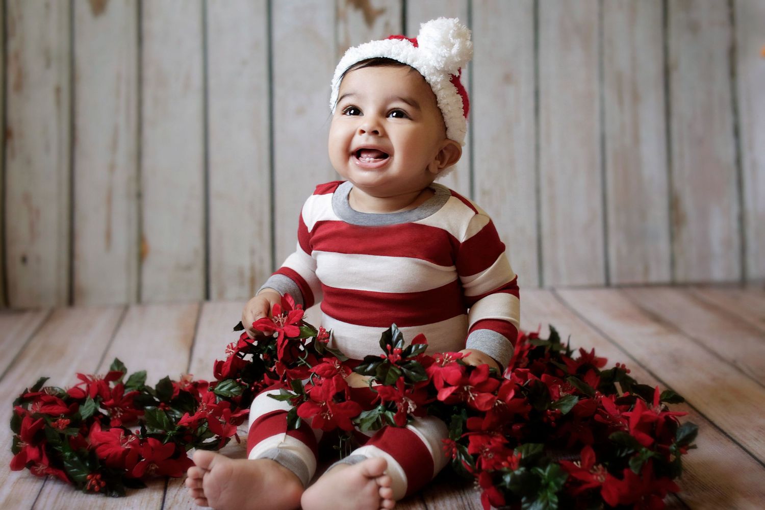 Smiling baby in red striped outfit and white hat sitting on wood floor surrounded by red holiday flowers.